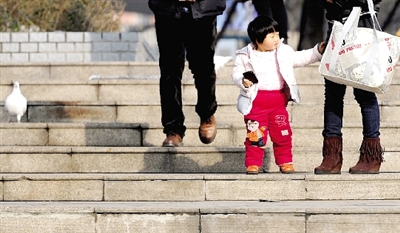 昨天中午，一女孩拉著家長的手在紫荊山公園曬太陽，氣溫升高，孩子的棉衣解開了扣子。